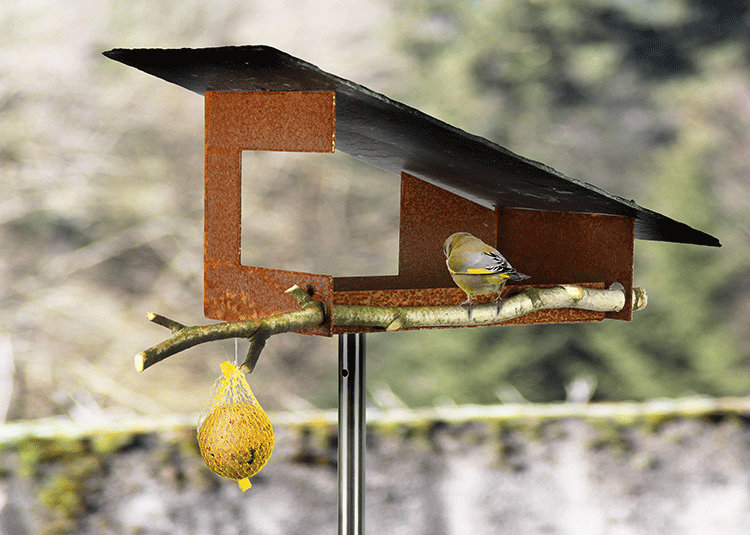 Detail van het voederhuisje van roest en staal, met tak en zittende vogel.