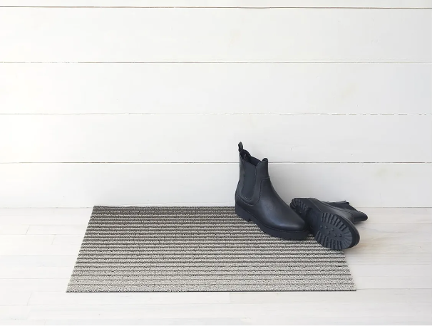 Deurmat Domino Oak van Chilewich, gestreept in beige-grijs, met laarzen ernaast