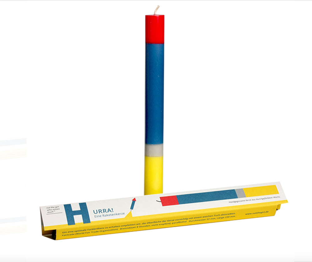 Speciale kerst-kaarsen van Schumacher Rabba, staand naast verpakking, in rood, blauw en geel.