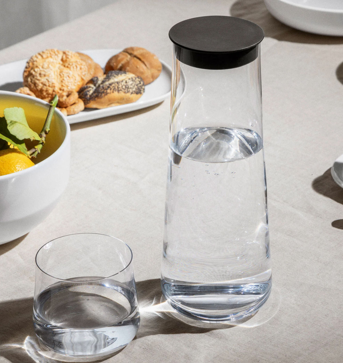 Alessi Eugenia karaf van glas met zwarte dop, gevuld met water naast een glas