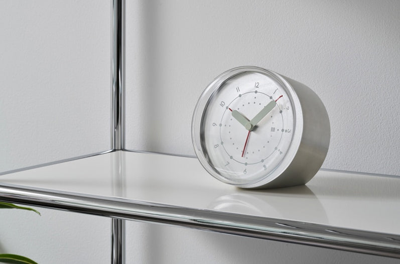 Alessi Ento analoge wekker, aluminium met witte wijzerplaat, vooraanzicht op een schap