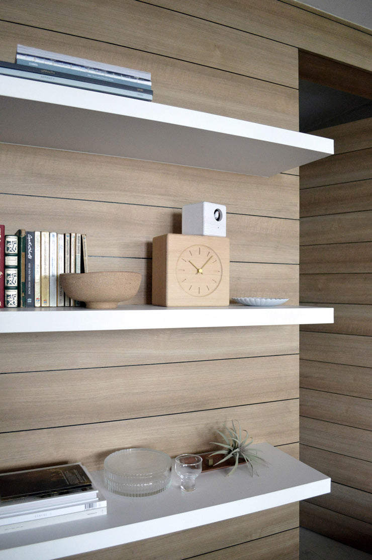 Lemnos koekoeksklok in hout en wit, op een wandplank tussen boeken en een schaal