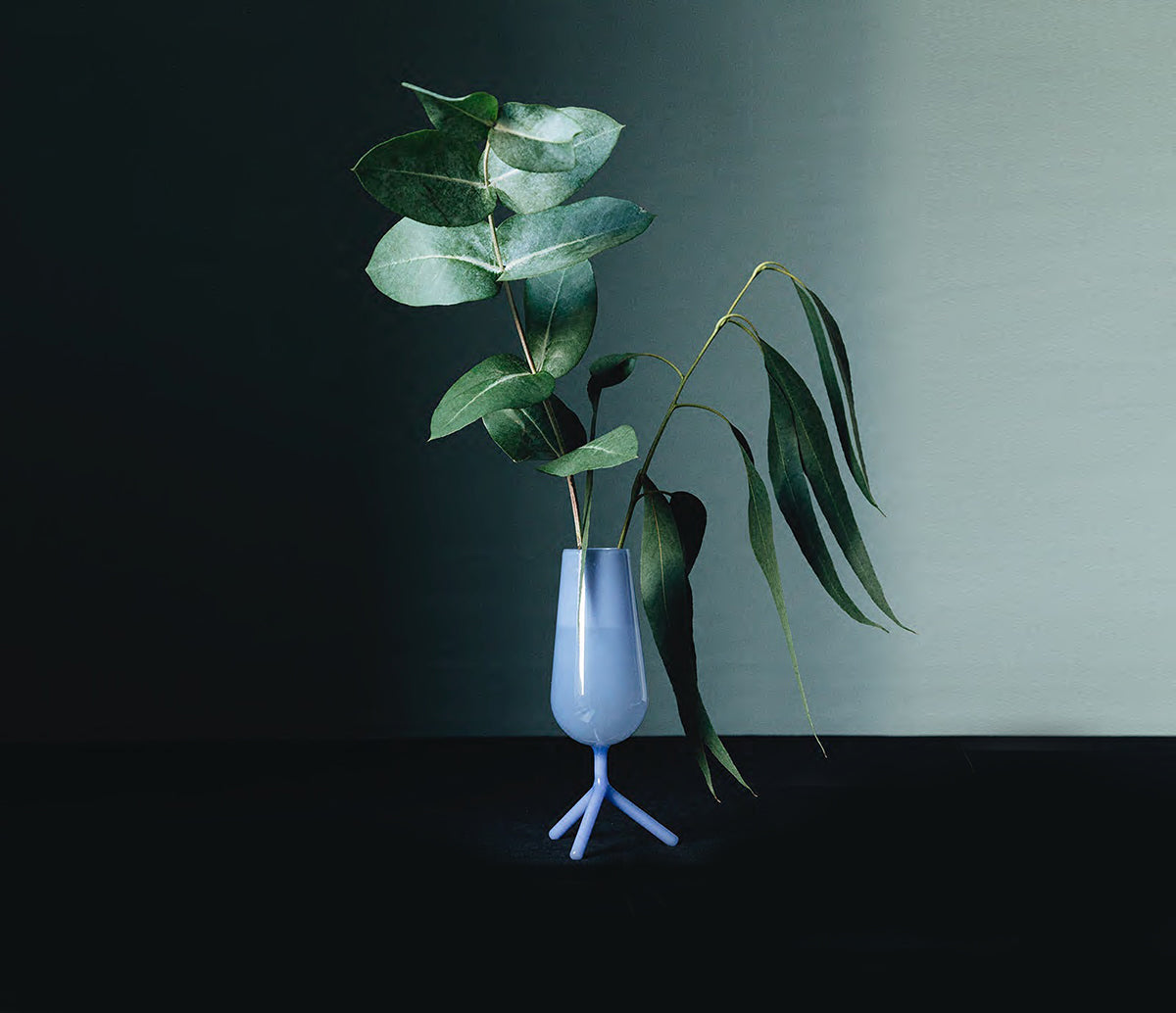 Kleine vaas in lavendelblauw glas op driepootvoet, met groene bladtakken